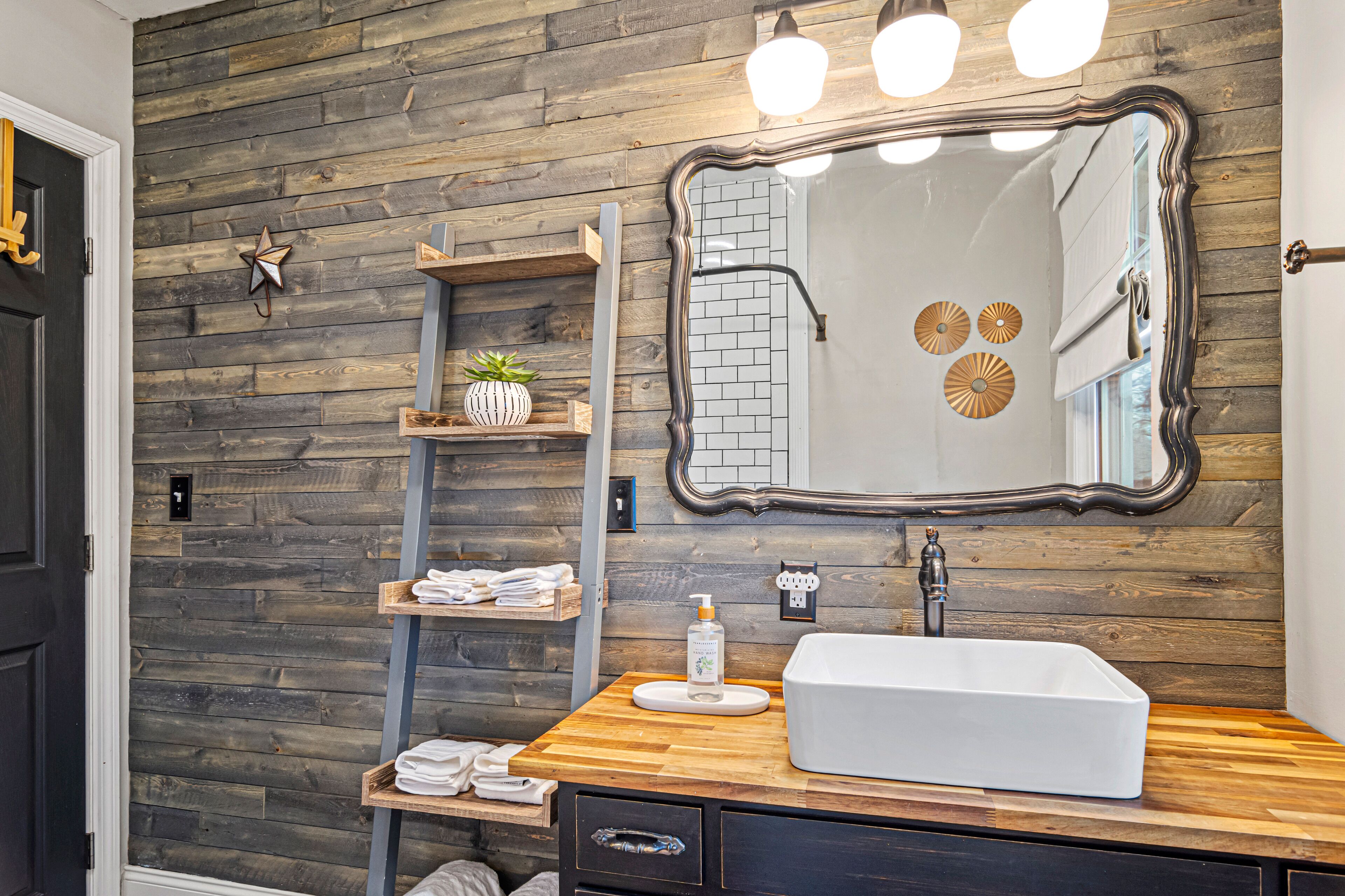 Un baño con pared de madera, un espejo grande, un lavamanos blanco y estantes con toallas y una planta.
