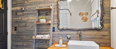 Un baño con pared de madera, un espejo grande, un lavamanos blanco y estantes con toallas y una planta.