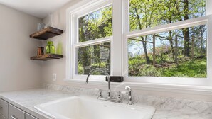 A kitchen with a large white sink, wooden shelves, and a window with a view of trees. - Farmhouse Writer's Retreat in Historic Shepherdstown (Shepherdstown)