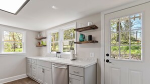 A kitchen with white cabinets, a marble countertop, and a window with a view of greenery. There are wooden shelves above the counter and a white door with a window. - Farmhouse Writer's Retreat in Historic Shepherdstown (Shepherdstown)