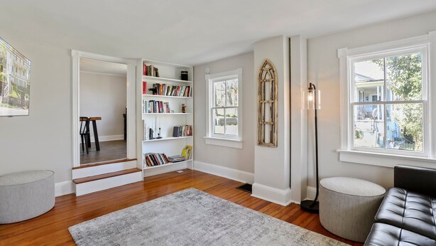 A spacious living room with hardwood flooring, a grey rug, a black leather sofa, a grey ottoman, a bookshelf, a tall floor lamp, and a window with a view of the outside. - Farmhouse Writer's Retreat in Historic Shepherdstown (Shepherdstown)