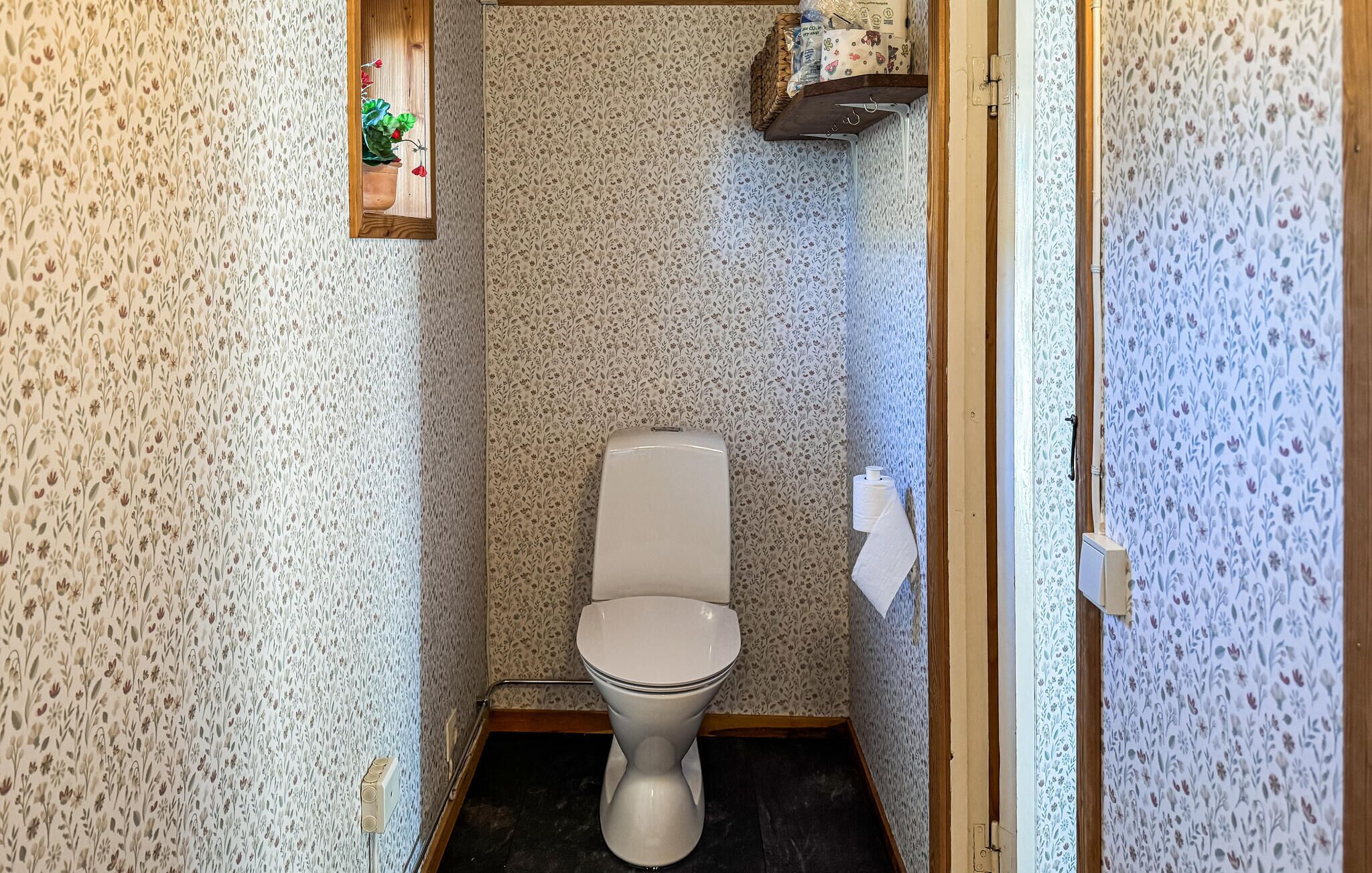 A small, narrow bathroom with floral-patterned wallpaper, a toilet, a hand dryer, and a small shelf with a potted plant.