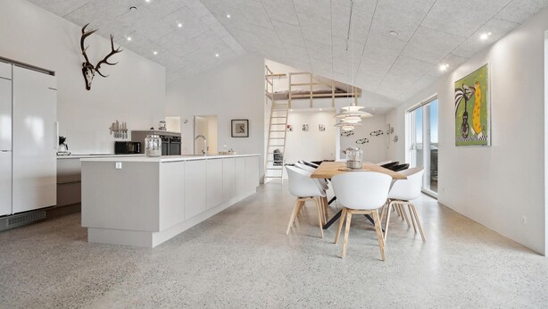 Una cocina moderna con una isla central, un área de comedor con una mesa de madera y sillas blancas, y una escalera que sube a un nivel superior.