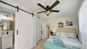 Un dormitorio con cama, ventilador de techo, mesita de noche y una puerta que da a otra habitación.