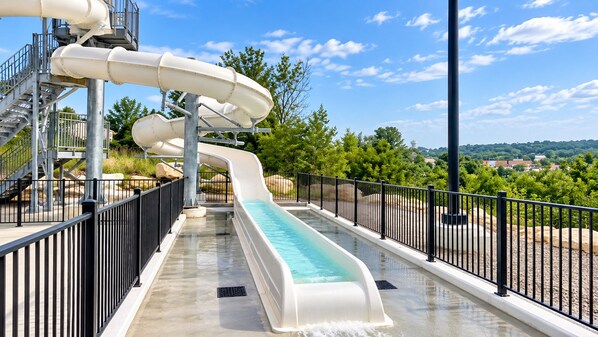 A water slide with a straight run and a pool at the end, surrounded by a metal fence and trees in the background.