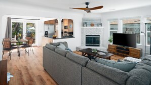 A modern living room with a grey sectional sofa, a dining area with a table and chairs, a kitchen with a built-in stove, and a fireplace. - Single-family PB house, 1 Block to Beach (San Diego)