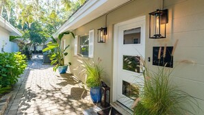 Un étroit sentier en briques mène à une maison aux murs recouverts de bardage blanc, agrémentée de plantes en pot et de lanternes fixées au mur.