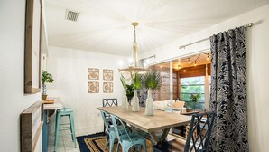 Un coin repas avec une table en bois, des chaises de couleur turquoise et un lustre. On trouve des plantes en pot sur la table et une fenêtre avec des rideaux.