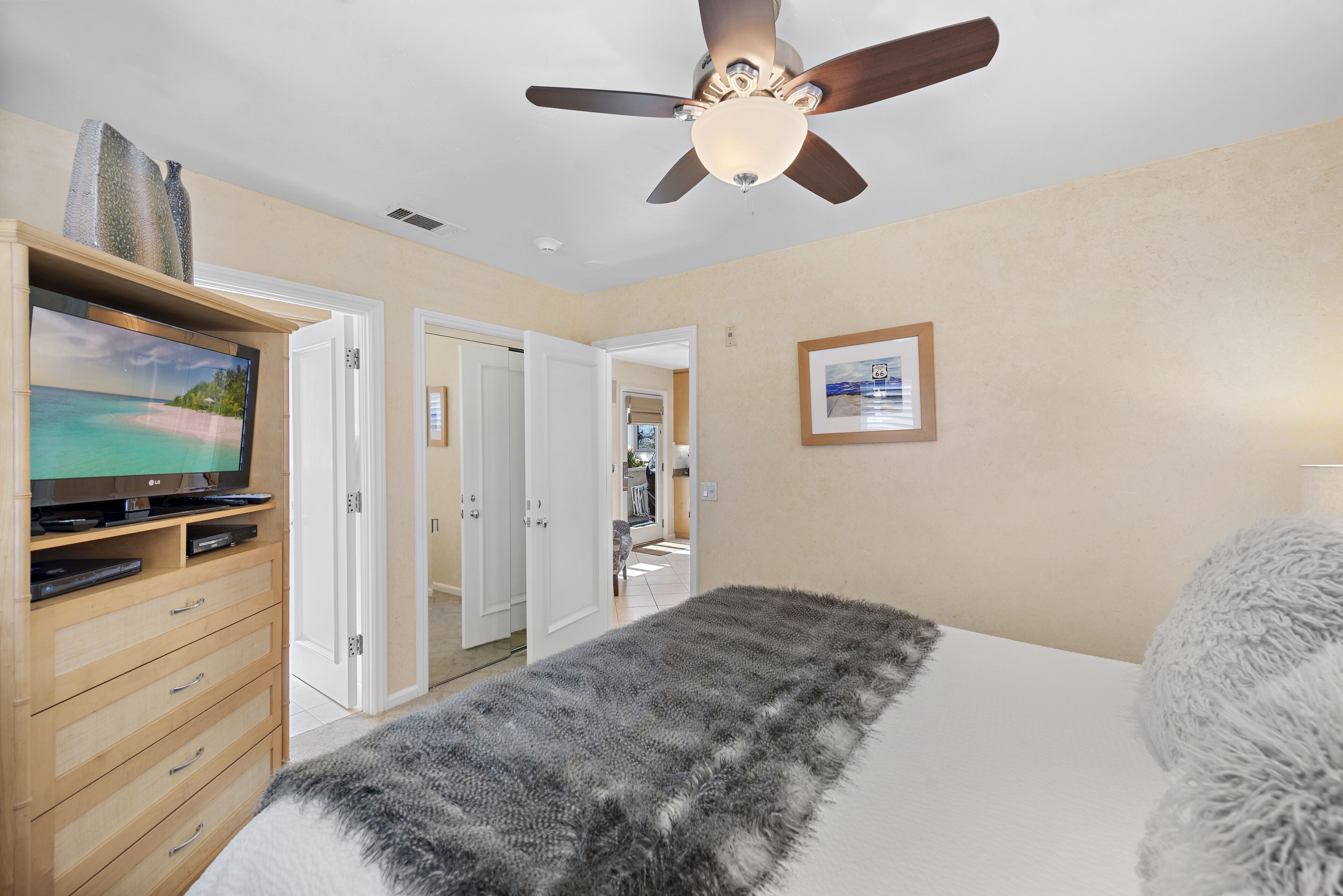 Un dormitorio con una cama, un televisor sobre un mueble de madera, un ventilador de techo y un cuadro en la pared.