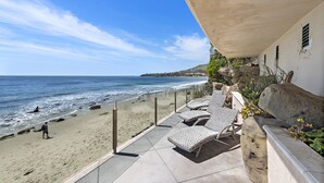 Une terrasse au bord de la plage, dotée de chaises longues, offrant une vue sur l’océan et s’ouvrant sur un paysage rocheux en arrière-plan.