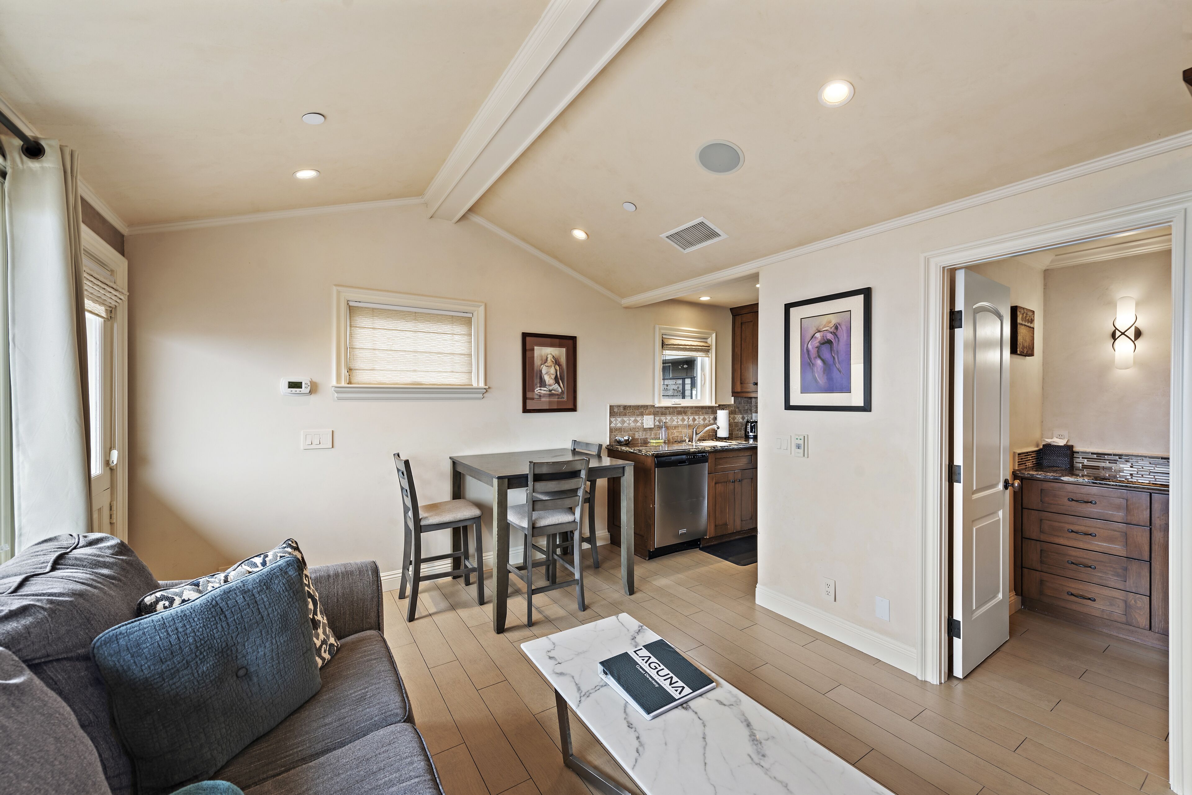 A spacious living area with a sofa, a dining table with chairs, a kitchen in the background, and a hallway leading to a bedroom.