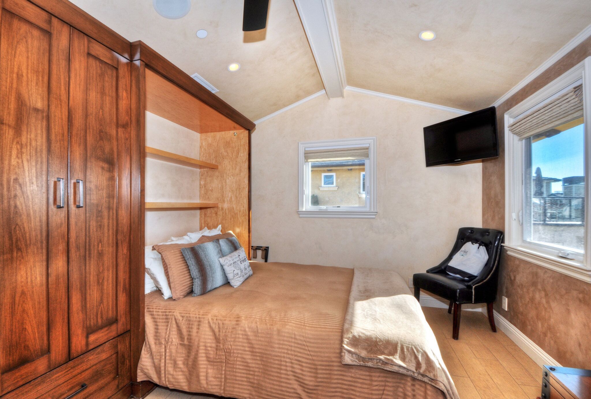 Un dormitorio con un armario grande de madera, una cama con muchos almohadones, un ventanal pequeño, un televisor montado en la pared y una silla.