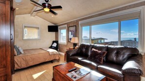 A spacious living room with a leather sofa, a wooden coffee table, and a ceiling fan. There is a bed with a window, a TV mounted on the wall, and a large window offering a view of the ocean. - Swim, bike, relax, explore Laguna Beach 3 Villa D'Arte (Laguna Beach)