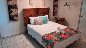 Un dormitorio con cabecera de madera, cama con ropa de cama blanca y almohadones de colores, un colgador de macramé en la pared y una puerta con perchero.