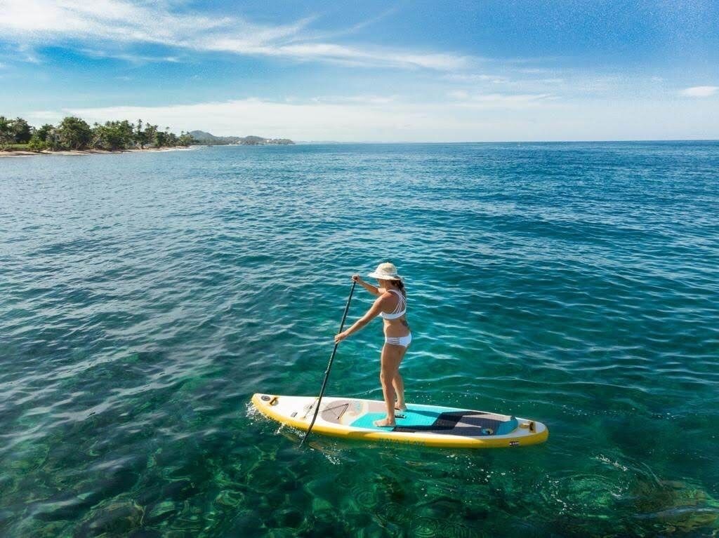 Eine Person steht auf einem Paddleboard auf ruhigem, blauem Meer, im Hintergrund eine Küstenlinie.