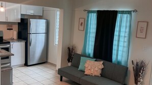 Una cocina compacta con refrigerador, horno y comedor. Un sofá gris con cojines se encuentra en la sala, y hay plantas decorativas y un cuadro en la pared.