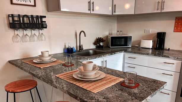 Una cocina con encimera de granito, armarios blancos, microondas, cafetera y dos taburetes.