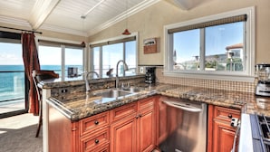 A kitchen with granite countertops, stainless steel appliances, and wooden cabinets. It features large windows with a view of the ocean and a balcony with a chair. - Ocean views steps to the water prime Laguna Beach 8 Villa Roma (Laguna Beach)