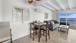 A spacious living area with a wooden dining table, comfortable seating, a ceiling fan, and large windows offering a view of the ocean.
