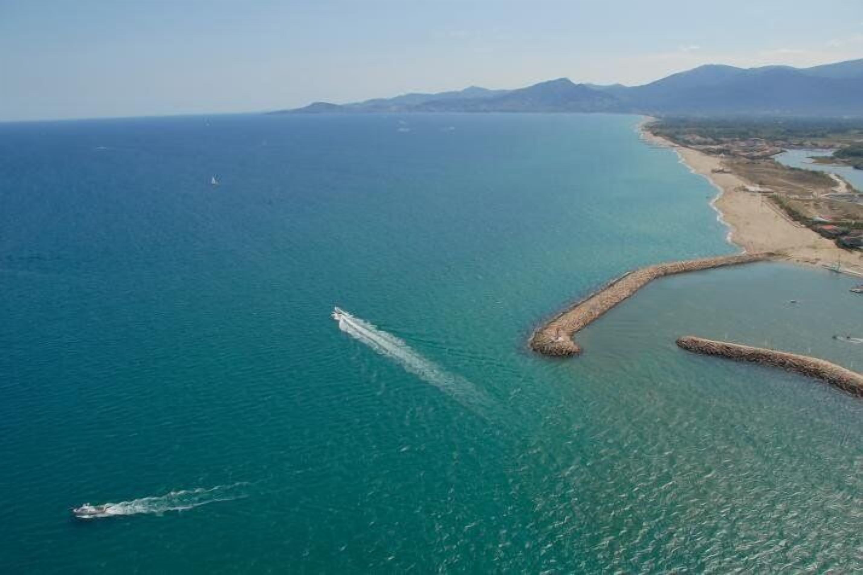 An aerial view of a coastal area with a clear blue sea, a sandy beach, and a breakwater structure.