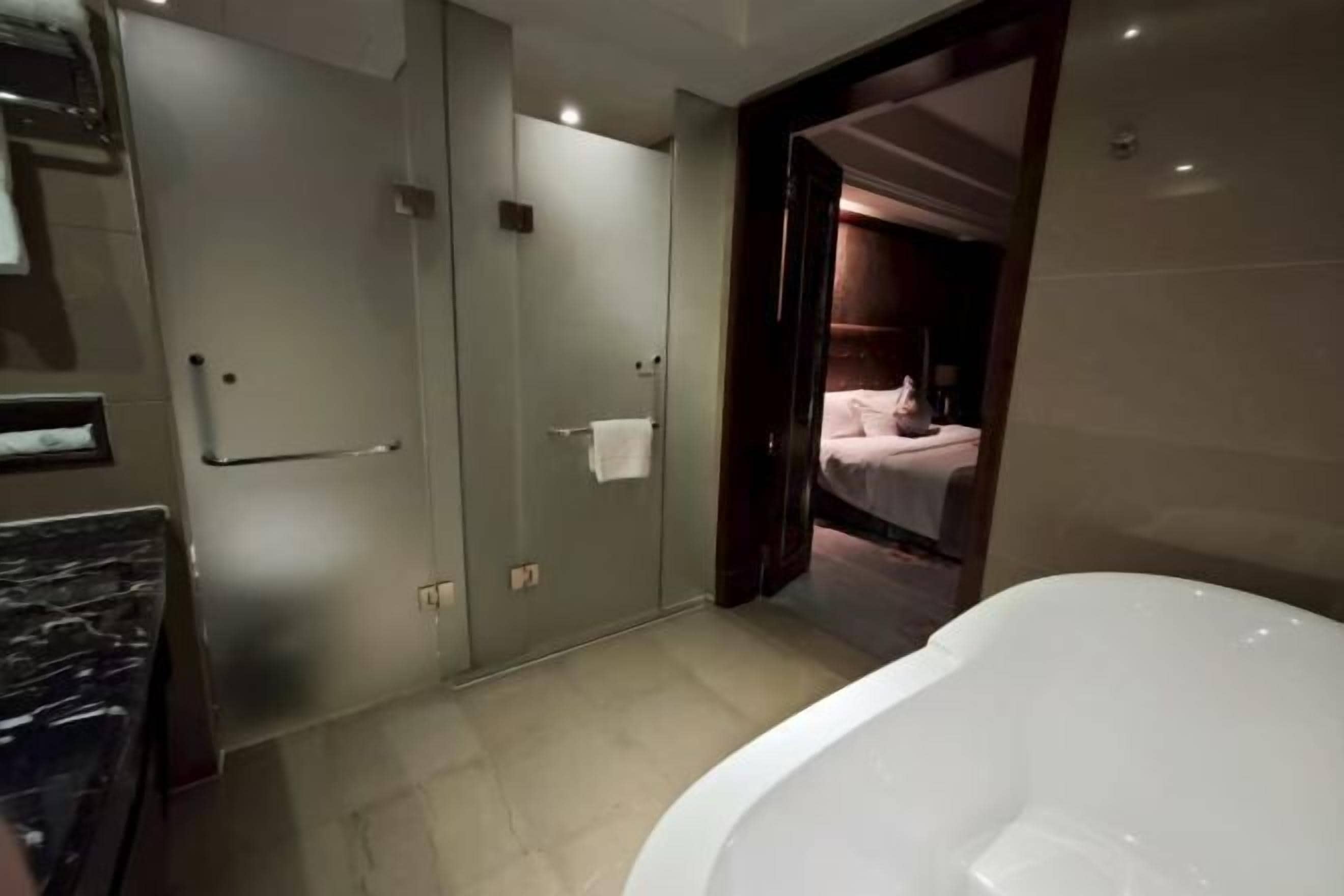 Un baño moderno con una bañera grande, un cerramiento de ducha de vidrio y una habitación visible con una cama.
