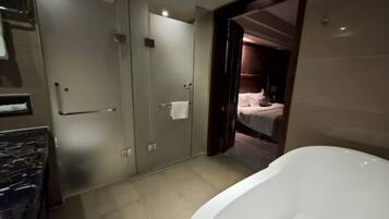 Un baño moderno con una bañera grande, un cerramiento de ducha de vidrio y una habitación visible con una cama.