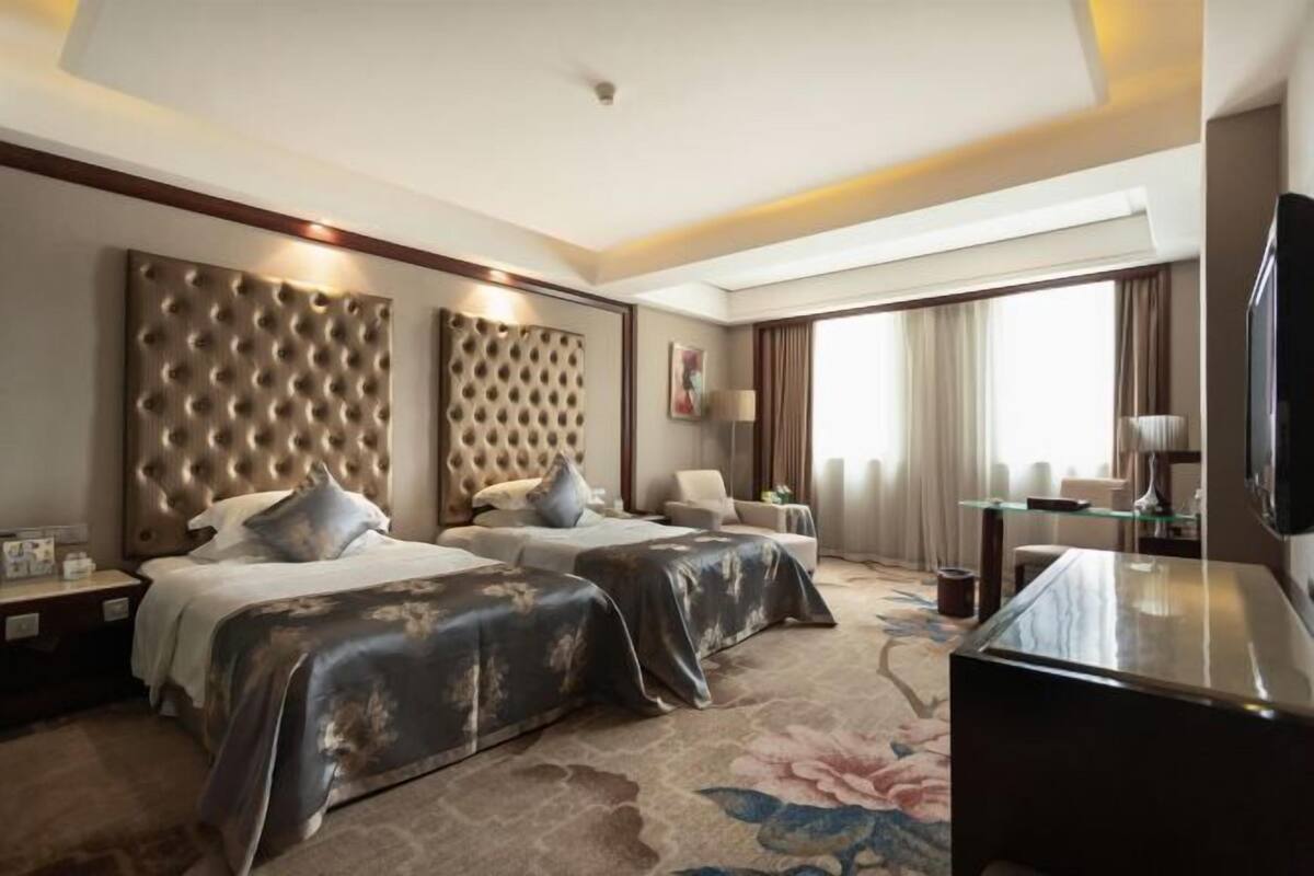 A hotel room with two beds, a large tufted headboard, a flat-screen TV, a glass coffee table, and a floral carpet.