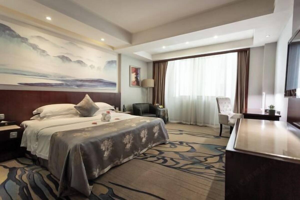 A hotel room with a large bed, a desk, a chair, a painting on the wall, and a window with curtains.