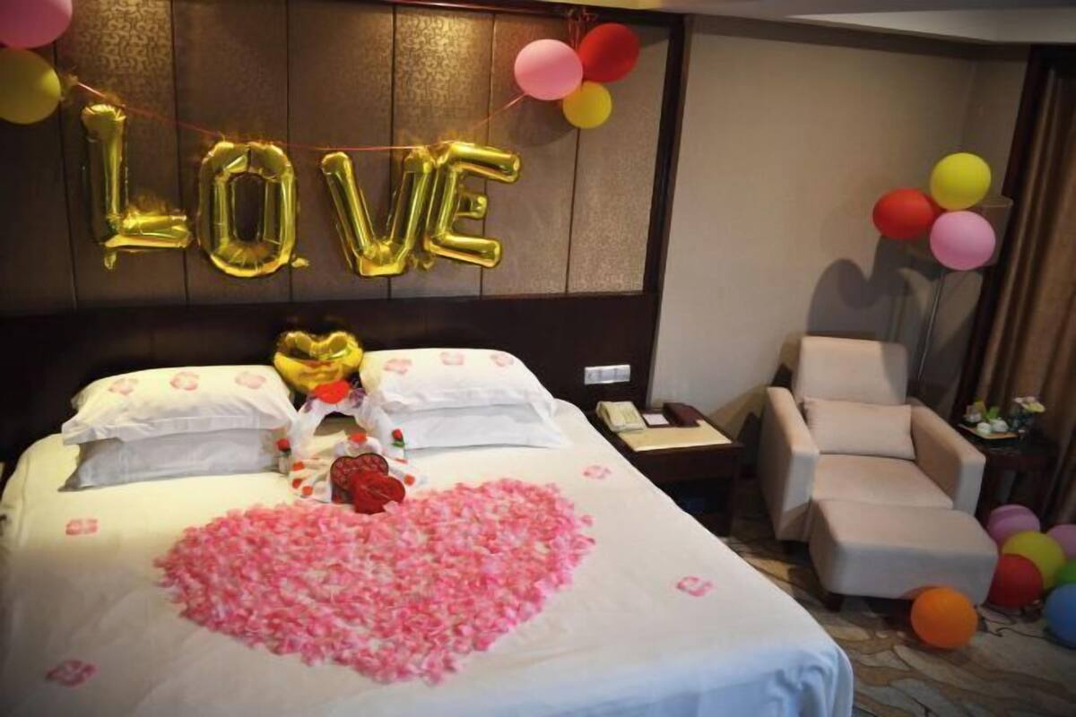 A hotel room with a bed, a heart-shaped arrangement of rose petals, a bouquet of flowers, and balloons spelling 'LOVE' on the wall.