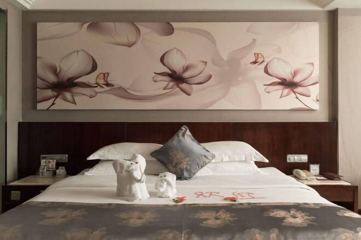 A hotel room with a large floral artwork on the wall, a neatly made bed with white linens, and a nightstand with a lamp and a phone.