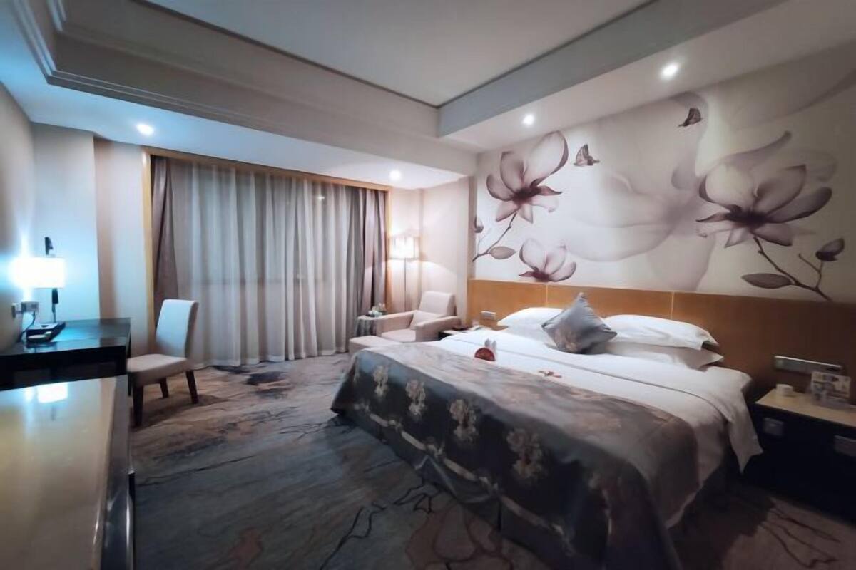 A hotel room with a large bed, a desk with a lamp, a chair, and a window with curtains.