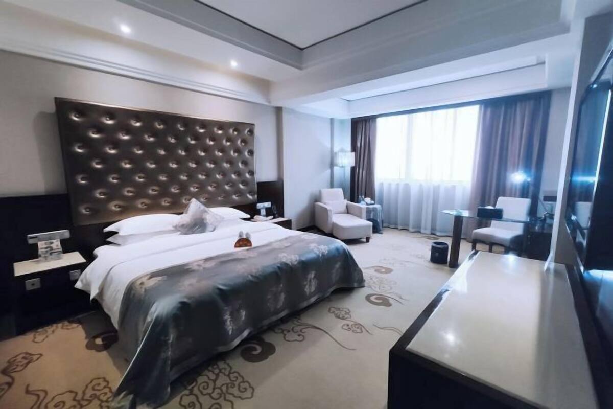 A modern hotel room with a large bed, a bedside table, a desk, and a chair.
