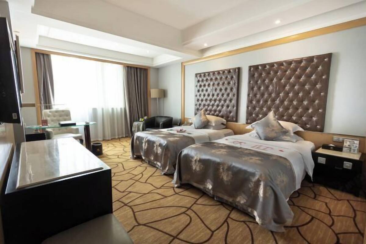 A hotel room with two beds, a TV, a desk, and a large window with curtains.
