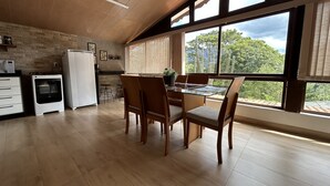 Une cuisine spacieuse avec un plancher en bois, un coin repas avec une table en verre et des chaises en bois, et de grandes fenêtres offrant une vue sur les arbres et un ciel nuageux.