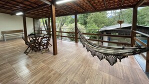 Une pièce spacieuse avec un plancher en bois, un hamac, une table en bois avec des chaises, et une vue sur la verdure à l’extérieur.