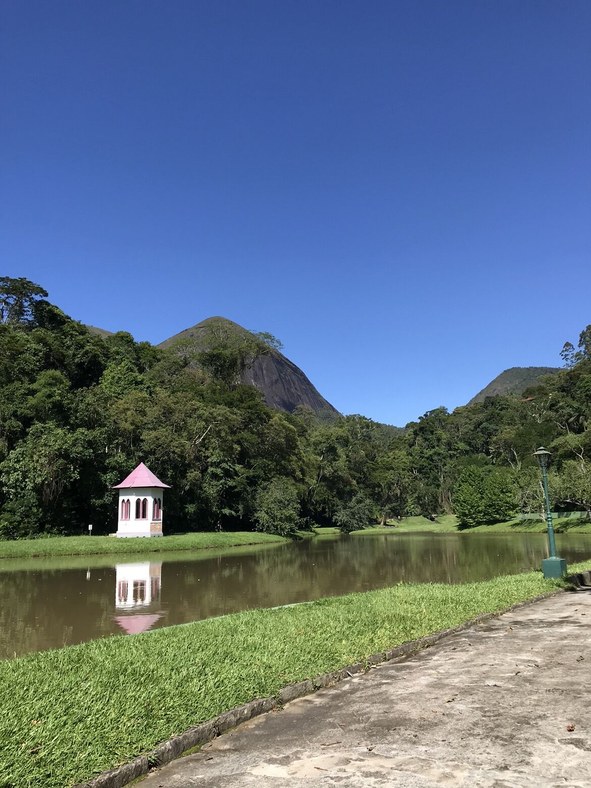 A tranquil lake with a small gazebo and a lamp post, surrounded by lush greenery and hills.