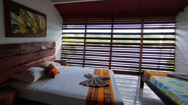 A bedroom with a wooden headboard, a bed with a white sheet and a decorative pillow, a bedside table, and a window with horizontal blinds. - Reserva Natural Yavary Tucano (Leticia)