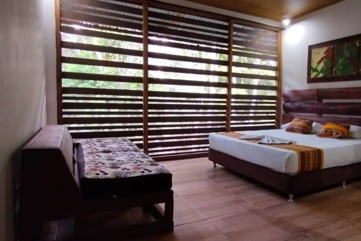 Une chambre à coucher avec un lit, un banc, une grande fenêtre avec des stores et un tableau accroché au mur.