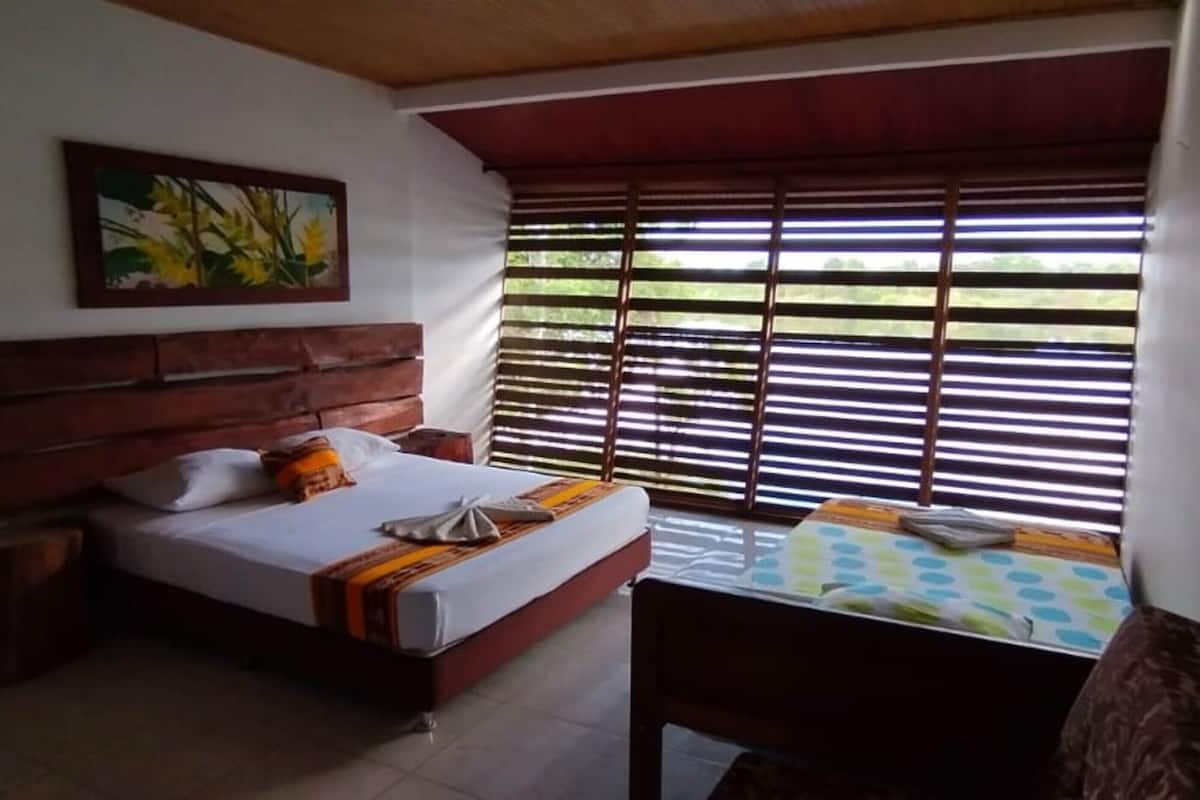 Une chambre à coucher avec une tête de lit en bois, un lit recouvert de draps blancs, une petite table avec une nappe à carreaux et une fenêtre avec des stores horizontaux.