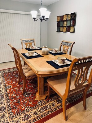 En spisestue med et træbord, stole, en lysekrone, en vase og et tæppe med et mønster.