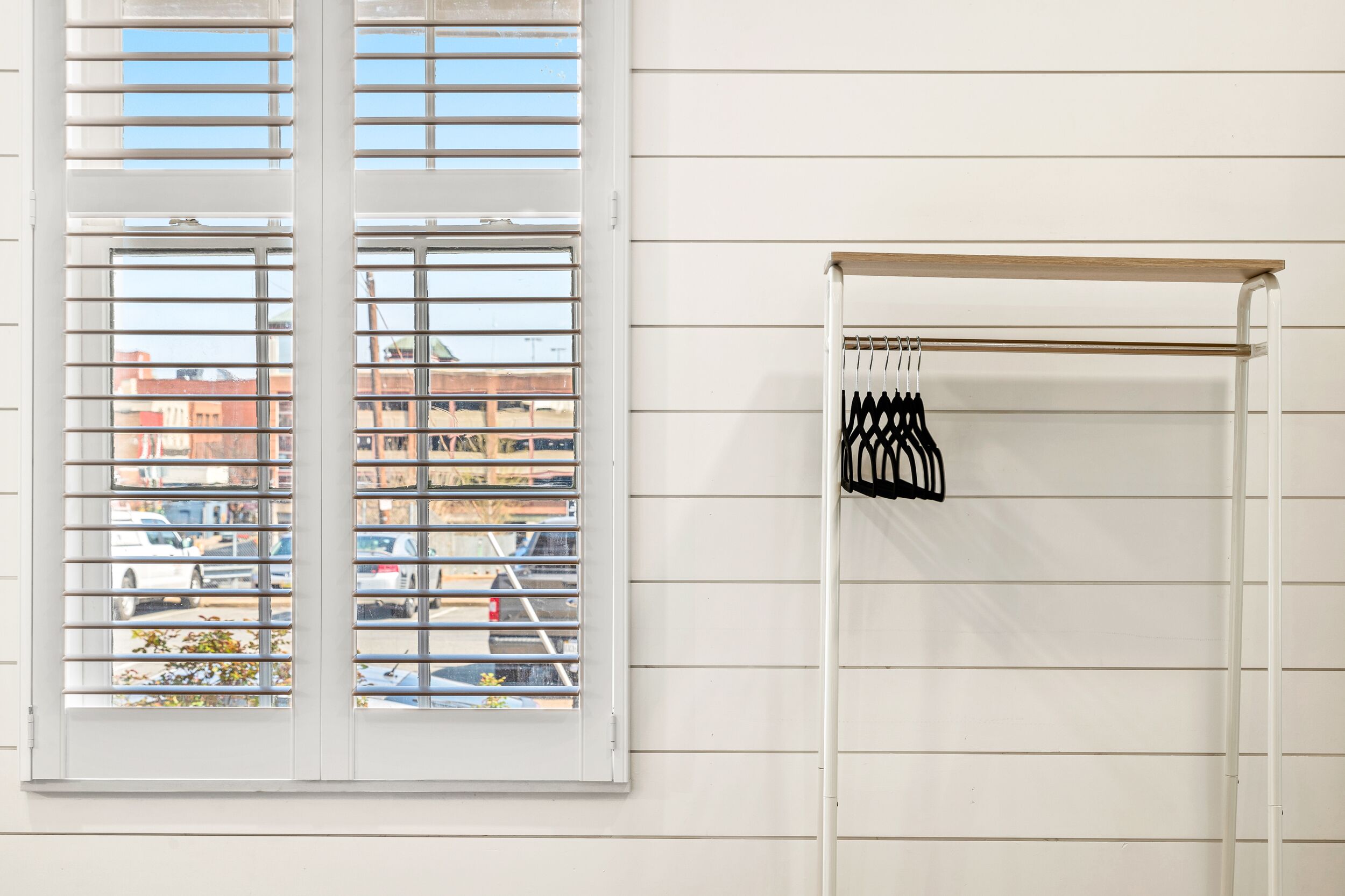 A room with a window with horizontal blinds, a white wall, and a clothes drying rack with black hangers.