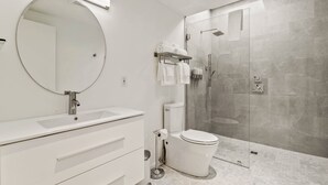 Un baño moderno con un espejo redondo, tocador blanco, inodoro y cabina de ducha con mampara de vidrio.