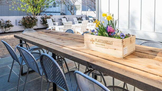 A wooden table with metal chairs, a wooden crate with flowers, and a white fence in an outdoor setting.
