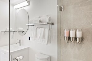 Une salle de bain moderne avec une douche à parois de verre, un lavabo blanc, des toilettes et un porte-serviettes où sont rangées des serviettes pliées avec soin. Trois distributeurs de savon sont fixés au mur.