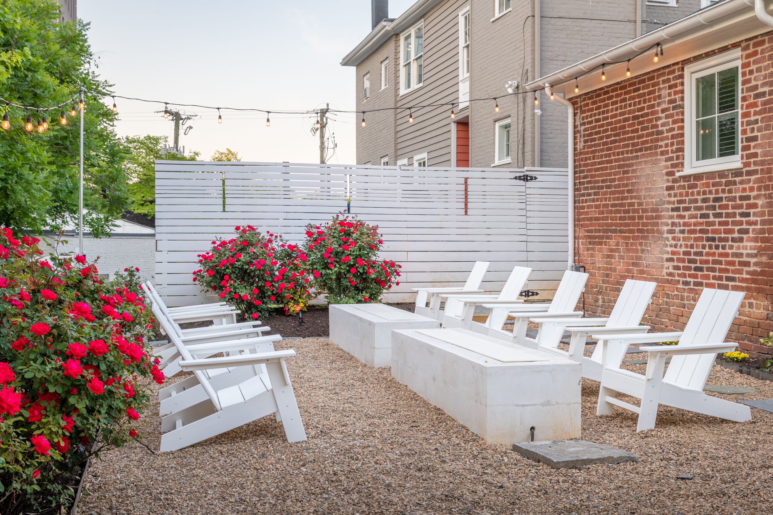 Un espace patio avec des chaises Adirondack blanches et une table en béton, entouré de gravier et de fleurs rouges.