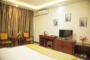 A hotel room with a bed, a desk with a television, a chair, a small table with a vase of flowers, and a coat rack.