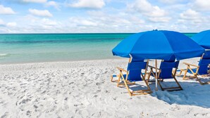 Una spiaggia con sabbia bianca e acqua cristallina color turchese. Si vedono ombrelloni blu e sdraio di legno.