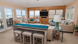 A spacious living room with a large TV, wooden paneling, a dining area with a glass table, and a view of the beach. - Ocean Views, Den, Community Pools, Close to Beach access, 103, Brigadune-Oceanfront (Corolla)