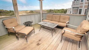 A covered outdoor seating area with a wooden deck, cushioned chairs, and a metal table. - Ocean Views, Den, Community Pools, Close to Beach access, 103, Brigadune-Oceanfront (Corolla)