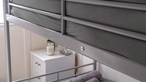 Una cama litera con ropa de cama gris, una mesita de noche blanca pequeña con una vela y una toalla gris en el suelo.